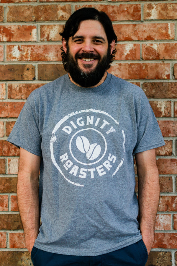 Man wearing a grey t-shirt with 'Dignity Roasters' logo against a brick wall.